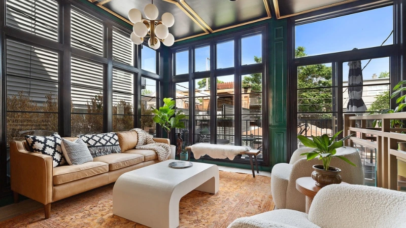 Sunlit enclosed porch with a tan leather sofa, white boucle chairs, and floor-to-ceiling windows in dark green trim in Chicago.