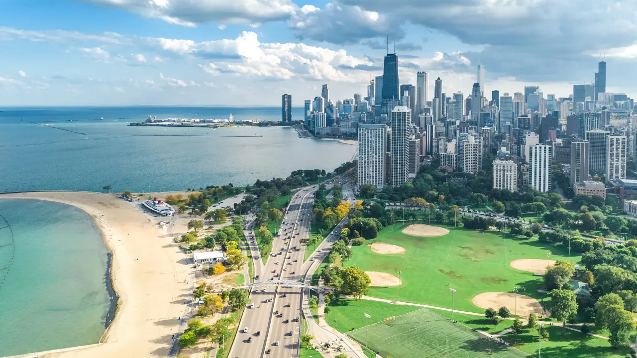 Aerial view of Chicago's lakefront, showing Lake Shore Drive, sandy beaches, green parkland, and the downtown skyline.