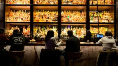 People sit at a bar with shelves of illuminated liquor bottles behind them.