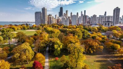 An aerial view shows Chicago’s skyline rising behind colorful fall trees in a large city park.