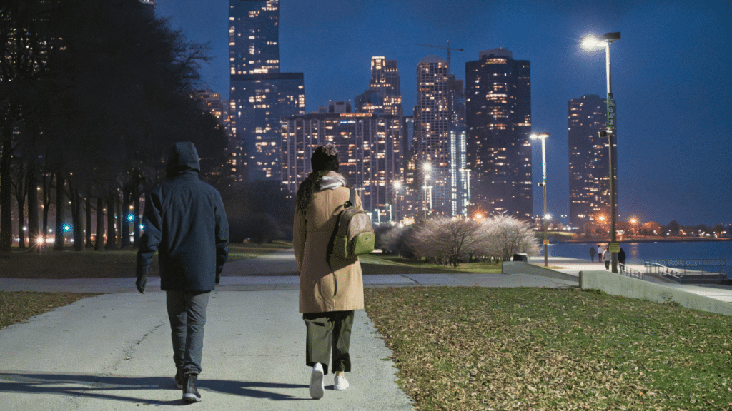 Two people walking along a lakefront path at night with the illuminated Chicago skyline in the background.