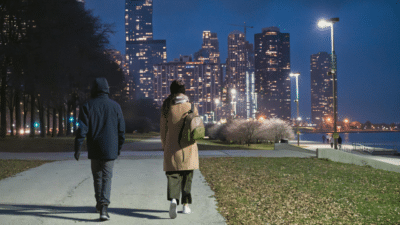 Two people walking along a lakefront path at night with the illuminated Chicago skyline in the background.